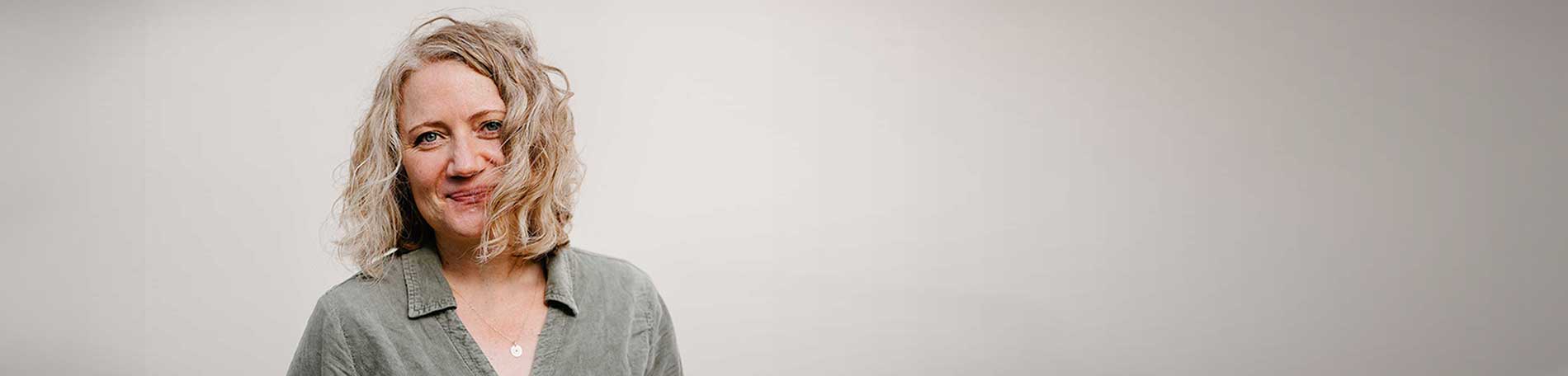 Ein Porträt von Sylvia Paproth. Sie hat kinnlanges, gewelltes, helles Haar, trägt eine olivgrüne Bluse mit einer dünnen goldenen Halskette und einem kleinen, kreisrunden Anhänger. Sie lächelt in die Kamera. 